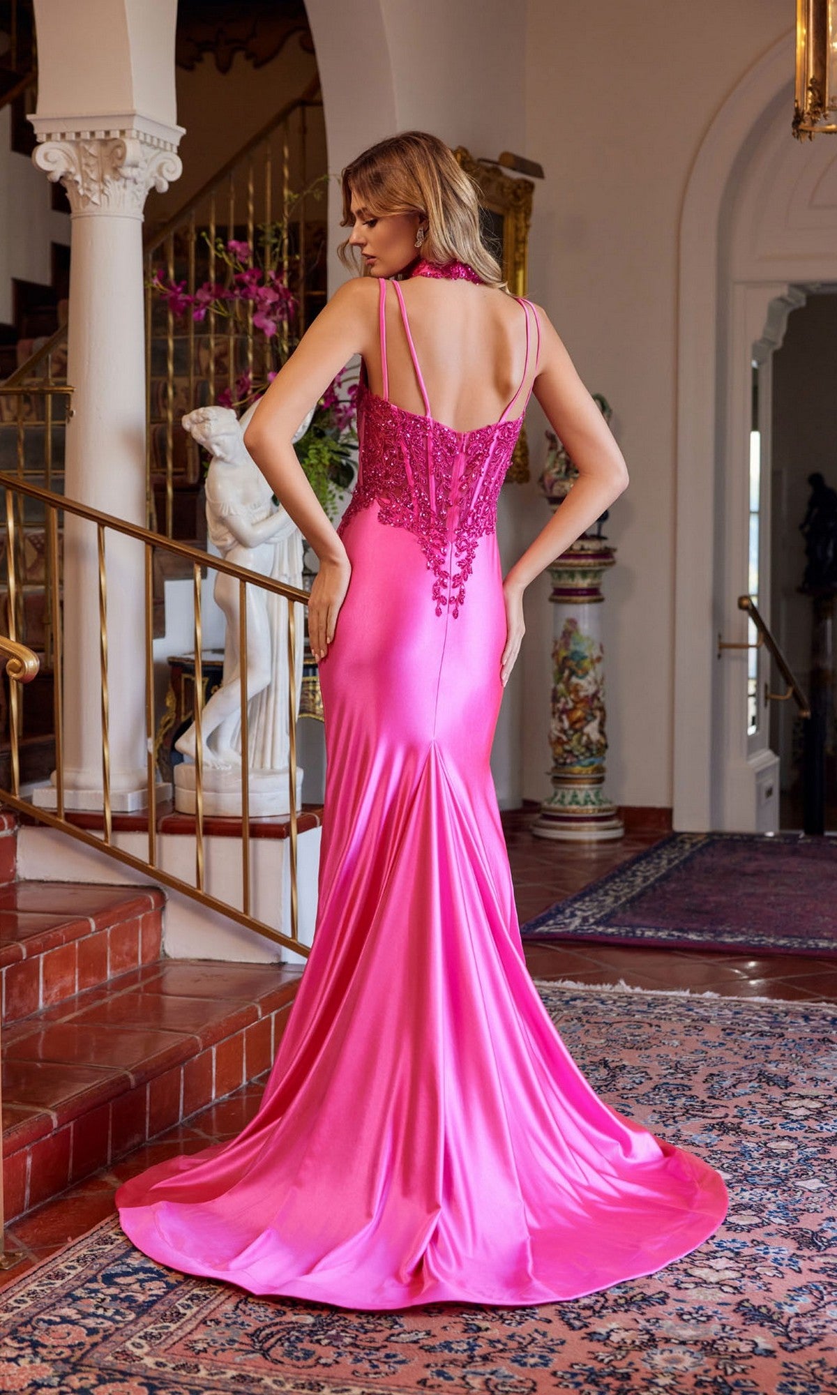 A woman stands indoors on a patterned rug, wearing the Juliet JT2628L long prom dress—floor-length, fitted, bright pink with a sweetheart neckline, beaded bodice, and long train—facing away from the camera.